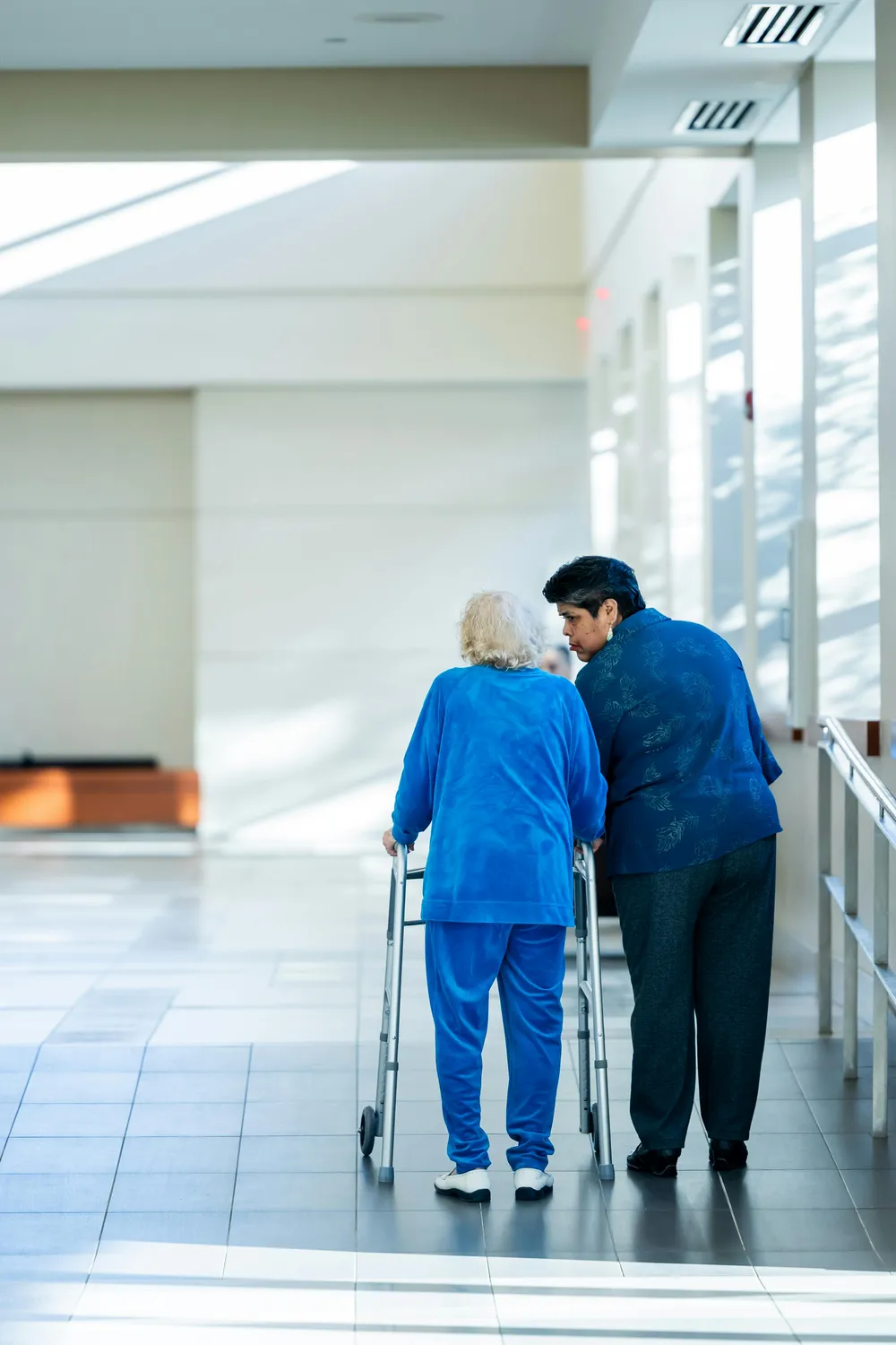 FamilyCare Leipzig UG – Pflegeleistungen für alle Altersgruppen – Eine Pflegekraft oder Krankenschwester unterstützt einen älteren Menschen mit einem Rollator in einem hell erleuchteten Flur