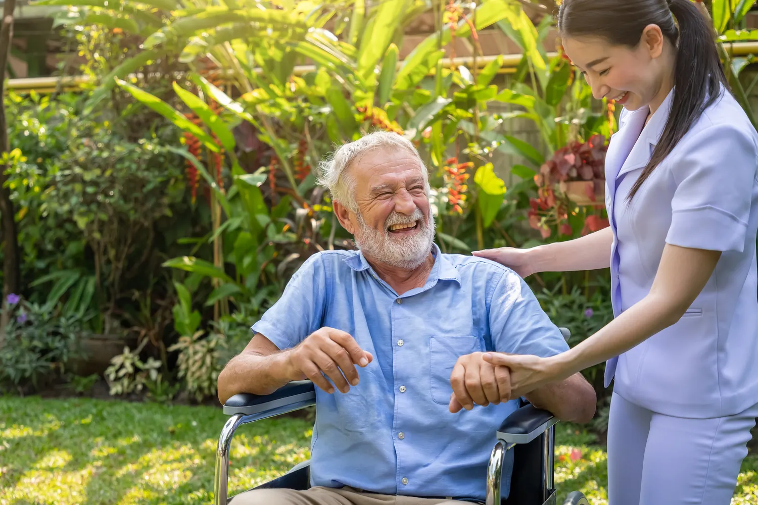 FamilyCare Leipzig UG – Pflegeleistungen für alle Altersgruppen – Fröhliche Pflegekraft hält lachenden älteren Mann im Rollstuhl im Garten des Pflegeheims